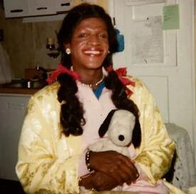 A photograph of Marsha P Johnson. Her hair is shoulder length, dark brown, and lightly curled, tied in pigtails with red bows. She wears red lipstick, and a yellow robe. She is smiling, posed for the photograph, and holding a plush Snoopy doll.